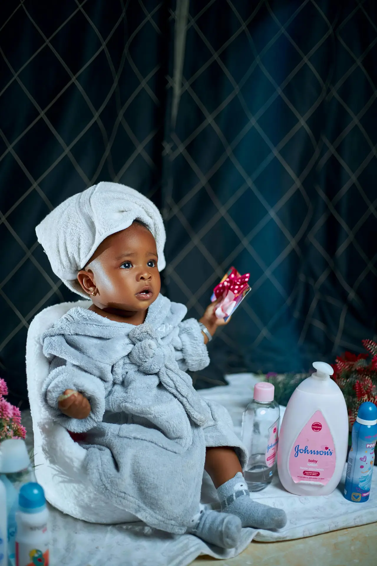 A cute toddler in a bathrobe holding a toy, surrounded by baby toiletries, sitting indoors.